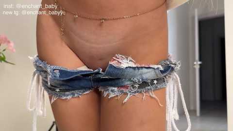 Media: Video of a light-skinned woman with a flat stomach wearing frayed, denim shorts pulled down, revealing her pubic area, in a modern, indoor setting with a pink flower in the background.