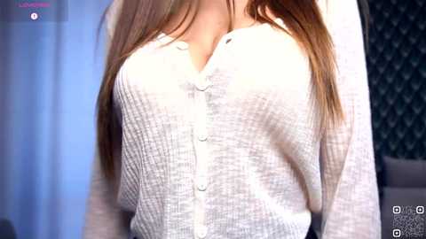 A video of a woman with long, straight brown hair, wearing a white, button-up, ribbed-knit cardigan. The background features a blurred, dark patterned wall and a hint of light blue curtains.