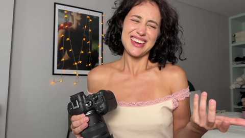 Video of a smiling, fair-skinned woman with curly black hair, wearing a beige off-shoulder top, holding a black camera, in a brightly lit room with a framed picture and string lights on the wall.