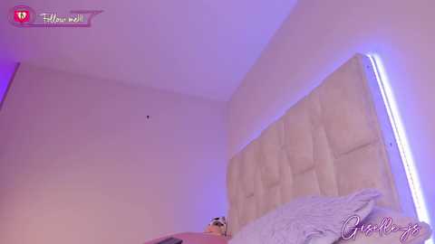 A video of a modern bedroom with a plush beige tufted headboard and purple LED lighting, featuring a plush white blanket and a small toy bear.