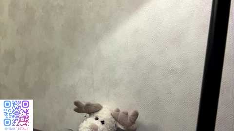 Video of a small, white plush reindeer toy with gray antlers and brown eyes, positioned on a textured, light gray wall. The image includes a QR code in the bottom left corner.