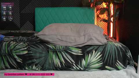 Video of a messy bed with a green pillow and a black floral duvet, against a dark, geometric-patterned wall. A lit candle and a phone display are visible in the background.