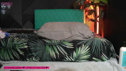 Video of a messy, unmade bed with a black and green leaf-patterned duvet, a gray pillow, and a teal headboard. Dark, geometric-patterned wall in the background.