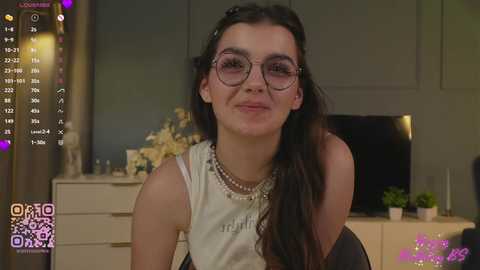Media: Video of a young woman with long dark hair, wearing glasses, a white tank top, and multiple pearl necklaces, seated in a cozy room with a TV, plants, and a dresser in the background.