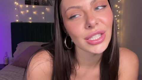 Media: Video of a young, fair-skinned woman with long brown hair, wearing silver hoop earrings, leaning forward with a playful, slightly puckered expression. Background features a bed with a dark headboard and string lights.