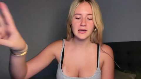 Media: Video of a young Caucasian woman with long blonde hair, wearing a low-cut gray tank top, holding her hand up, in a dimly lit room with dark furniture.