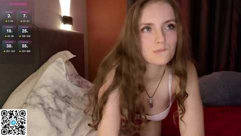 A video of a young, fair-skinned woman with long, wavy brown hair and a white tank top, lying on a bed with a marbled comforter, in a dimly lit room with a digital clock displaying 2:07 AM.