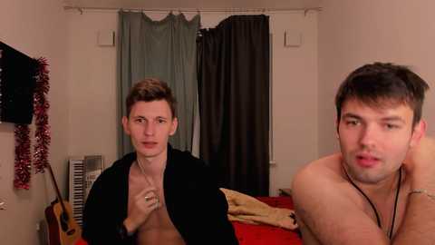 Video of two shirtless young men in a bedroom; one has a black jacket, the other a black necklace, both looking at each other.