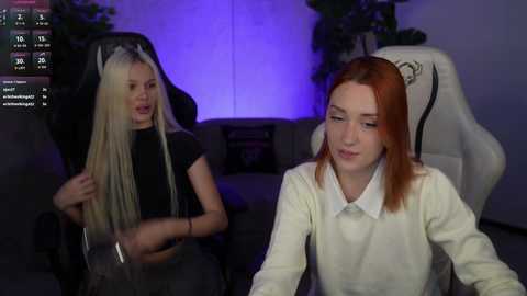 Video of two young women in a dimly lit room; one with long blonde hair in a black top, the other with red hair in a white sweater, sitting on white gaming chairs.