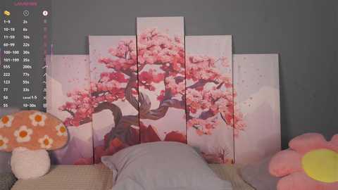 A video of a bedroom with a gray wall featuring a wall art of blooming cherry blossoms, a plush toy with a pink flower, and a pillow with a floral pattern.