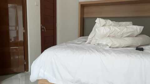 Media: Video of a minimalist bedroom with a neatly made bed covered in white sheets, a wooden headboard, and a closed wooden door on the left.
