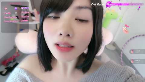 A close-up video of an East Asian woman with straight black hair, fair skin, and closed eyes, wearing a gray off-shoulder sweater. Background includes a computer screen with streaming chat.