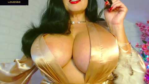 Media: Video of a curvy woman with long black hair and red lipstick, wearing a low-cut, shiny gold satin robe, revealing ample cleavage. Background shows a white brick wall and pink flowers.