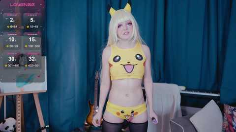 Video of a young woman with fair skin, wearing a Pikachu-themed crop top and shorts, with blonde hair, in a bedroom with blue curtains, a Pikachu plush toy, and a gaming setup.