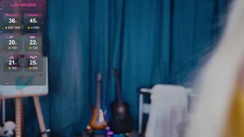 A video shows a dimly lit room with a blue curtain backdrop. A guitar leans against a white stand, and a small screen displays a song's lyrics. The scene is slightly blurred, creating a dreamy ambiance.