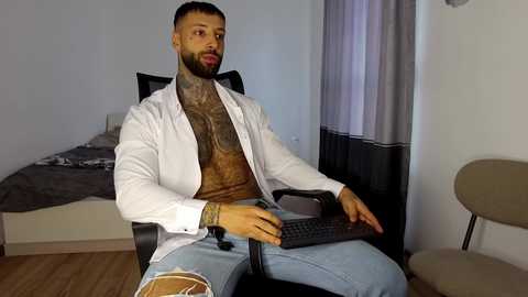 Video of a muscular, bearded man with a large tattoo on his chest, wearing a white shirt and jeans, sitting in a black chair, typing on a laptop, in a simple, modern bedroom with a bed and beige curtains.