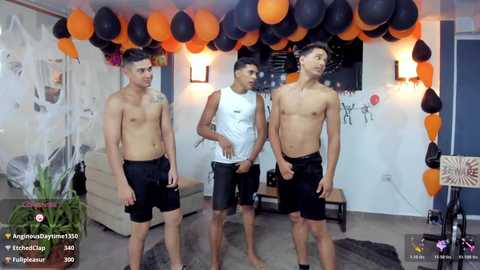 Three shirtless, muscular young men in black shorts pose indoors, surrounded by orange and black balloons, Halloween decorations, and a white couch.