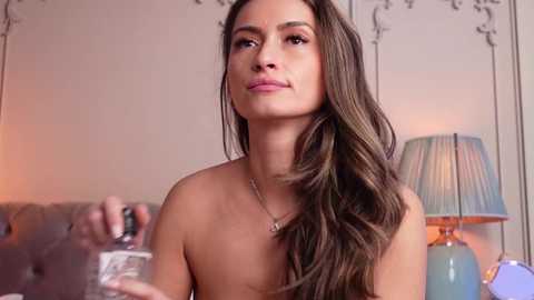 Media: Video of a topless woman with long, wavy brown hair, holding a small bottle of liquid, in a softly lit, elegant room with a lamp and ornate wallpaper.