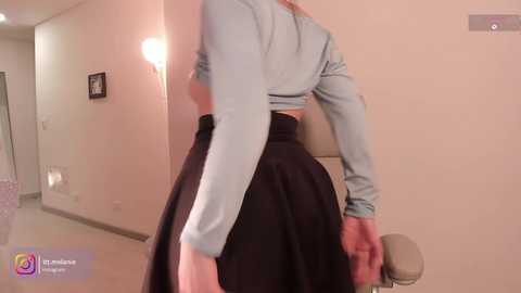 Video of a woman in a light blue long-sleeve crop top, black high-waisted skirt, and beige heels, walking down a hallway with beige walls and a lamp.