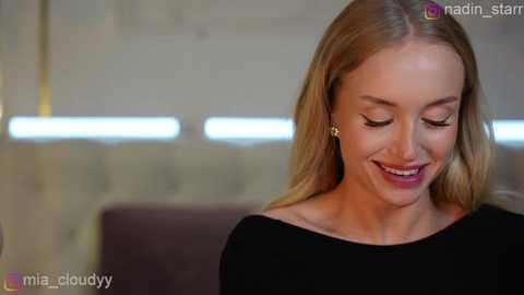 Video of a smiling, fair-skinned blonde woman with long hair, wearing a black top, against a blurred, modern indoor setting with bright lights.