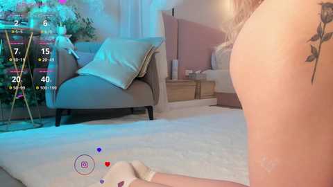 A video captures a nude woman with fair skin, standing with her back to the camera, in a cozy bedroom with a gray armchair, pink walls, and a plush carpet.