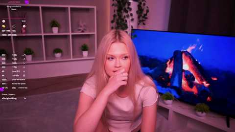 Video of a young woman with long blonde hair, light skin, and a medium build, wearing a beige shirt, sitting in a modern living room with a TV displaying a virtual fireplace.