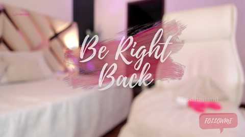 Video of a modern bedroom with a white bed, pillows, and a white armchair. The background is soft pink. Text reads \"Be Right Back\" in white cursive. A pink hairbrush is on the floor.