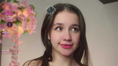 Video of a young girl with long brown hair, a gray hair clip, and pink lips, smiling. Background features a pink cherry blossom branch and a blurred, beige wall.