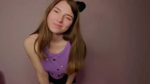 Media: Video of a young woman with light skin and long brown hair, wearing a purple tank top, black hairbands, and a slight smile. Background is plain, muted pink.