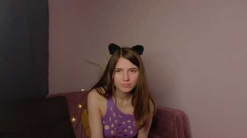 Media: A video of a young woman with long brown hair, wearing a purple polka-dot sleeveless top and black cat ear headband, sitting on a pink velvet couch against a pink wall.