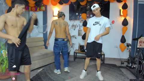 Video of three shirtless young men in athletic shorts, one in a backwards cap, standing in a dimly lit room adorned with orange and black balloons, posters, and a staircase.