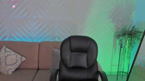 A video of a modern living room with a black leather gaming chair, beige sectional sofa, geometric-patterned wall, green and blue ambient lighting, and a tall potted plant.