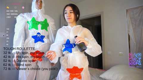 A video shows a young woman in a clear, star-patterned plastic jumpsuit, holding a spray bottle in a bedroom. The background features a bed, white walls, and a window with a curtain.