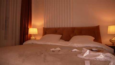 Video of a well-lit, cozy hotel room with a beige tufted headboard, two white pillows, a beige blanket, and two nightstands with lamps. A white lace bra is discarded on the bed.