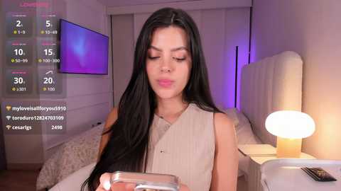 Video of a young Asian woman with long black hair, wearing a beige top, sitting on a bed in a modern bedroom with a lit lamp.