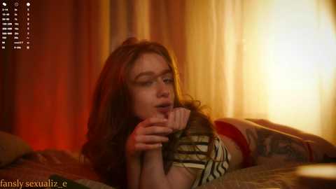 Media: Video of a young woman with long, wavy brown hair, lying on a bed, holding her finger to her mouth. Warm, golden-yellow light fills the room, creating a cozy atmosphere.