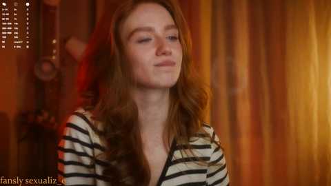 Media: A video of a young woman with long, wavy, light brown hair, wearing a black and white striped shirt. She has fair skin and a serene expression. The background features warm, orange and yellow curtains, creating a cozy atmosphere.