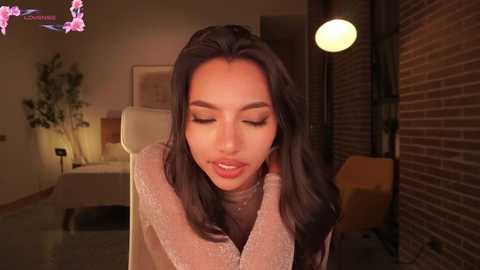 A video of a young Asian woman with long black hair, wearing a beige sweater, looking down with a serene expression. The background features a cozy, dimly-lit room with brick walls and a potted plant.