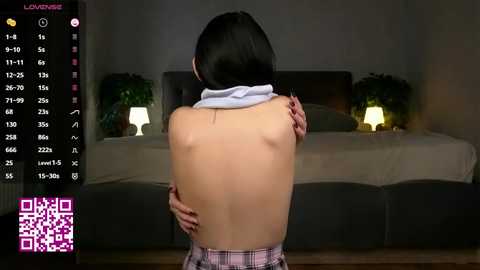 A video of a topless woman with fair skin and black hair, wearing only a plaid skirt, standing in a dimly lit bedroom with a bed and potted plants. A QR code is visible in the bottom left corner.
