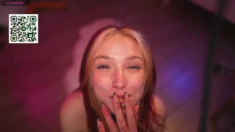 Video of a fair-skinned, blonde woman with a nose ring, covering her mouth with her fingers, set in a dimly-lit, blurred background with a QR code.
