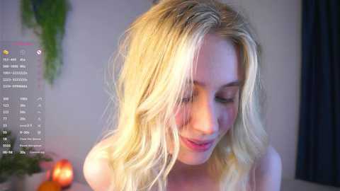 Video of a young woman with long, wavy blonde hair, fair skin, and a slender build, wearing a light-colored top, smiling softly. Background features a blurred indoor setting with soft lighting.
