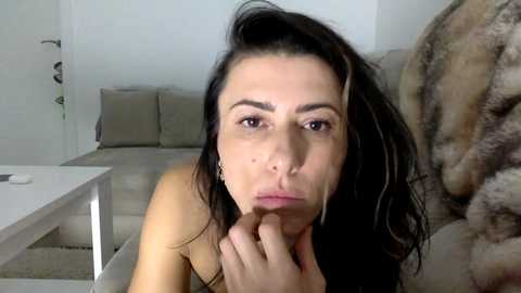 Media: A video of a young woman with long dark hair, light skin, and a contemplative expression, sitting in a beige living room with a white table and fur rug.