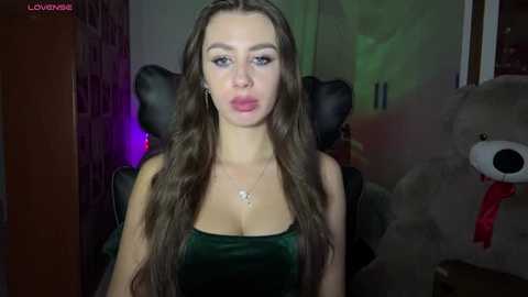 Video of a Caucasian woman with long, wavy brown hair, wearing a dark green velvet dress, standing in a dimly lit room with a plush teddy bear and a green wall.