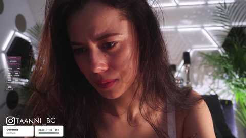 A video of a woman with long, dark hair, wearing a white top, in a modern, high-tech room with hexagonal lights and green plants, looking distressed.