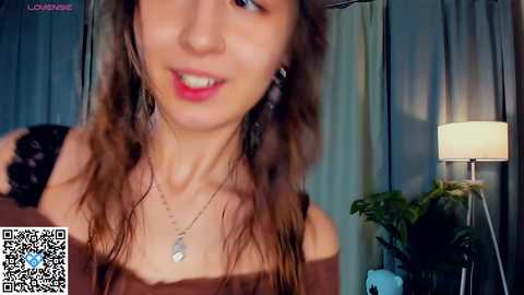 Media: A close-up video of a young woman with long brown hair, smiling with red lipstick, wearing a brown top, silver necklace, and standing in a dimly lit room with a green plant and a lamp.