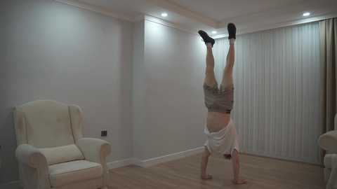 A video of a person doing a handstand in a minimalist living room with white walls, wooden floors, and beige furniture. The individual is wearing a white t-shirt and shorts, with legs raised and arms extended.