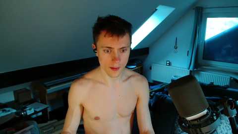 Video of a young, shirtless, fair-skinned, slim Caucasian man with short dark hair, sitting in a cluttered attic bedroom with sloped ceilings and skylights. Background includes a messy room with various objects, a desk, and a radiator.