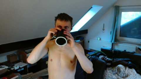 Video of a shirtless young man with short dark hair, holding a camera, in a messy, dimly lit attic room with sloped ceilings, a bed, and cluttered surfaces.
