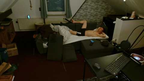 A video of a messy living room with a shirtless man sleeping on a couch, surrounded by a laptop, remote, and scattered items on the floor.