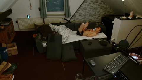 Video of a messy, dimly lit attic room with a man lying on a sofa, surrounded by boxes, a computer, and a cluttered desk.